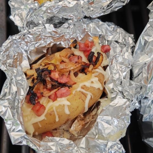 Fertige Grillkartoffel mit geschmolzenem Käse, Röstzwiebeln und Schinkenwürfeln in Alufolie