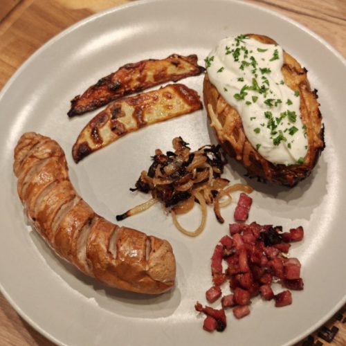 Servierte Grillkartoffel mit Sour Cream, Grillwurst, Schinkenwürfeln und Röstzwiebeln auf Teller